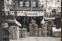 Epiciers à l'entrée de leur boutique, devant laquelle sont exposés des fûts de bière, de «miso»  (pâte de haricots de soja fermentée), de saké et du charbon de bois, Tôkyô, Japon, 1926-1927, (Autochrome, 9 x 12 cm), 
Roger Dumas, Département des Hauts-de-Seine, musée Albert-Kahn, Archives de la Planète, A 68 627 X